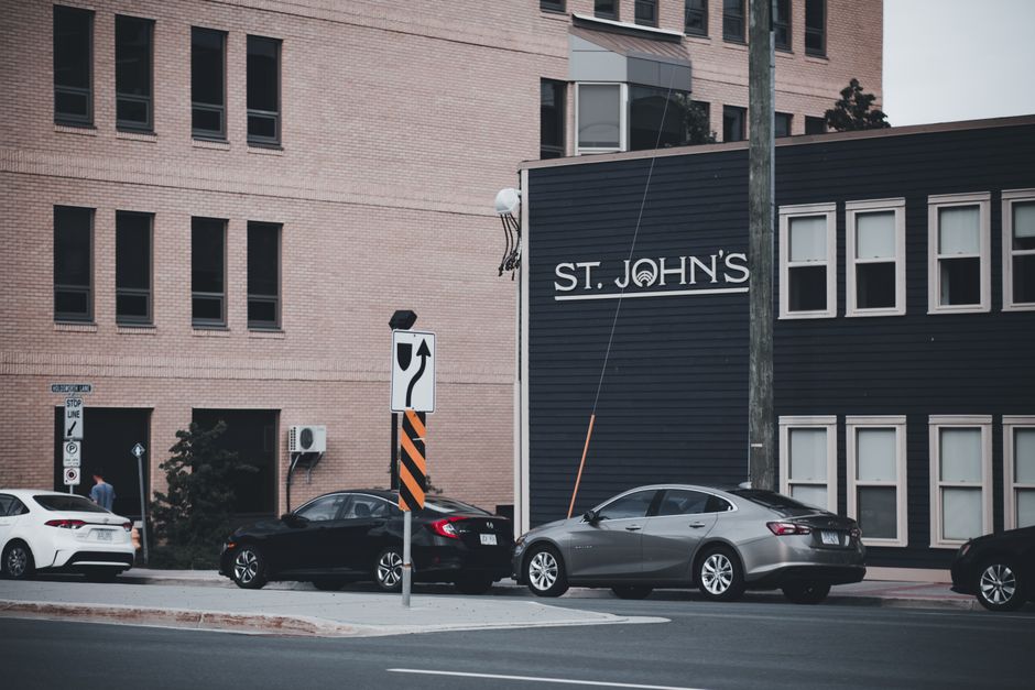 A city street scene shows a black and a silver sedan parked in front of a modern, multi-storey brick building with multiple windows. To the right, there is a dark-colored, horizontally paneled commercial or residential structure with six evenly spaced windows. A street pole with a white and black directional sign indicating a left turn is positioned in front of the vehicles. The background includes a tree and an external air conditioning unit attached to the brick building. The street surface appears clean and well-maintained, with a curb separating the road from the sidewalk. The overall atmosphere is typical of an urban environment, conveying a scene where private vehicle parking is common, supporting the context of alternative waste handling and private rubbish collection services often associated with urban property management, such as that provided by Waste Disposal St John's Wood.