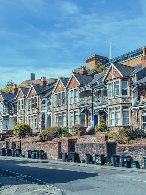 A row of Victorian-style terraced houses positioned on an inclined street, featuring intricate white iron lacework balconies and decorative gable ends painted in shades of red brick and cream. The properties are elevated above the pavement, accessible via small front gardens enclosed by low brick walls with steps leading up to each entrance. The street in the foreground is lined with multiple black wheelie bins placed along the curb, indicating ongoing waste collection activities. The background includes a modern building with glass windows and a rooftop structure, contrasting with the historic residential facades. The sky above is bright blue with scattered clouds, and the lighting suggests a clear daytime setting, emphasizing the architectural details and the tidy appearance of the waste bins and gardens. Waste Disposal St John's Wood offers private rubbish removal services suited for residences like these, facilitating alternative waste handling outside of local authority collection. The overall scene depicts a typical London residential street prepared for rubbish collection or disposal activities, with attention to cleanliness and orderly waste bin placement.