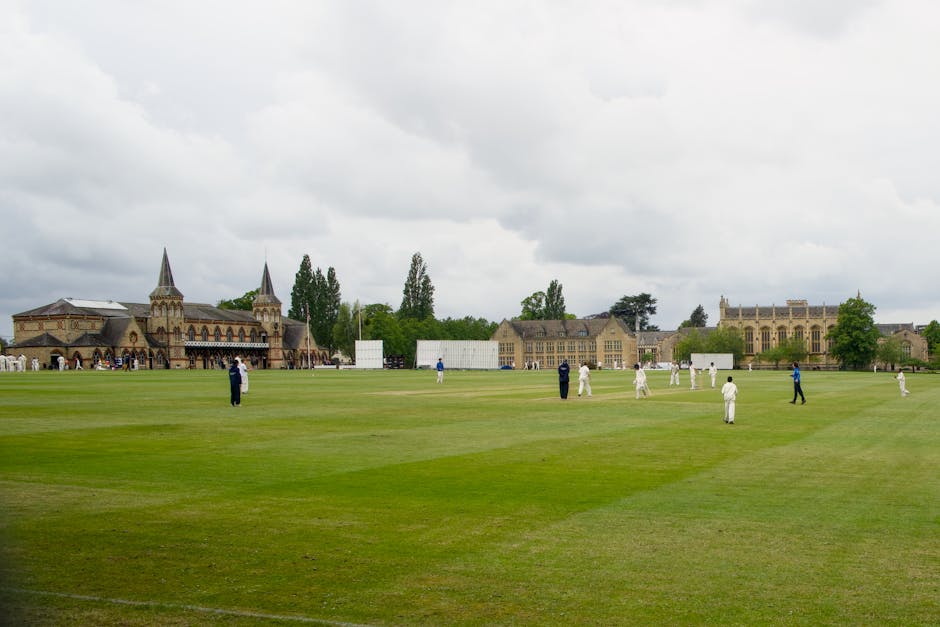 A wide view of a cricket ground with a well-maintained green pitch set in front of historic stone buildings and a church with tall, pointed spires, under a cloudy sky. Several players dressed in white cricket uniforms and some in darker clothing are scattered across the field, engaged in practice or game activities. In the background, there are trees and additional buildings, creating an environment typical of a private sports ground with surrounding structures that suggest a garden or park-like setting. The scene is outdoors, with natural diffuse lighting from the overcast sky, emphasizing the clarity of the grassy surface and the detailed texture of the stone architecture and the cricket players’ uniforms. The image reflects a peaceful, expansive environment associated with outdoor sports and leisure, aligning with the concept of private or alternative sport facility management, possibly related to independent grounds maintenance or event hosting, as seen in professional waste and disposal service contexts.