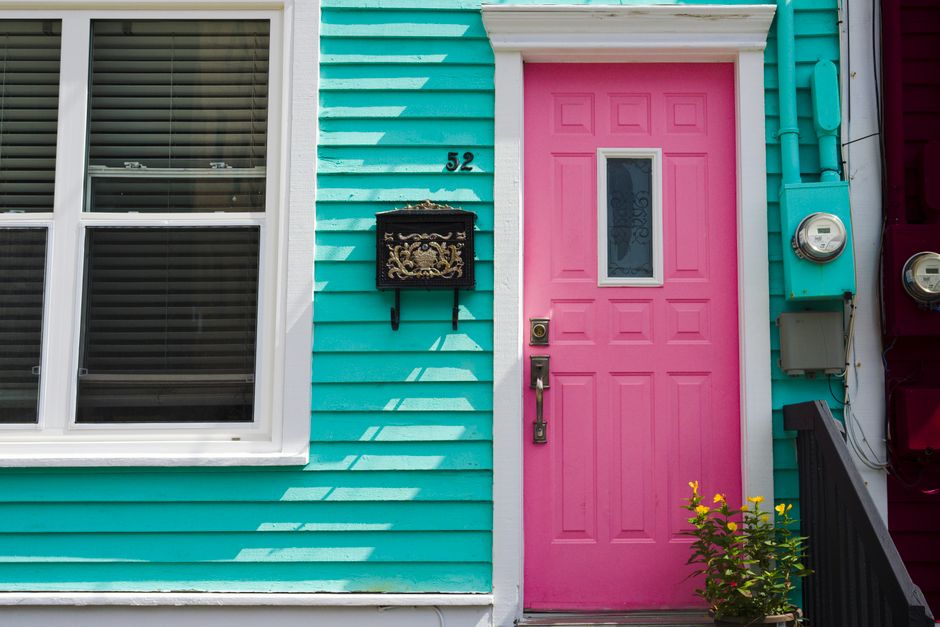 The image shows the front exterior of a house with brightly colored teal wooden cladding. A large white-framed window with horizontal blinds occupies the left side of the scene, while the focal point is a vibrant pink front door positioned slightly to the right. The door features a small vertical glass panel with decorative etched glass, a brass door handle, and a lock. To the left of the door, a black decorative mail collection box with ornate detailing is mounted on the wall, with the house number '52' displayed just above it. On the right side, part of a black staircase railing is visible leading up to the entrance, and a small green shrub with yellow flowers grows near the bottom of the steps. Adjacent to the door, there is a grey plastic electrical meter box and an outdoor electrical cable connected to a grey utility box, all mounted on the blue cladding. The environment appears to be well-lit with natural daylight, highlighting the contrasting colors and textures of the exterior materials, indicative of a residential property that may be serviced by private waste disposal or rubbish removal providers for NW8 homes.